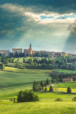 A picturesque hillside village in Italy, with a prominent church standing under a cloudy sky with sun rays breaking through, is surrounded by lush green fields and trees.
