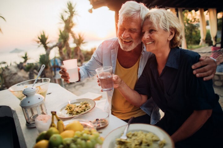 An elderly couple happily clinks their glasses while enjoying a meal outdoors in a tropical setting at sunset. A table with food and fruits is in front of them, embodying the spirit of travel and tourism as they savor the beauty of Greece.