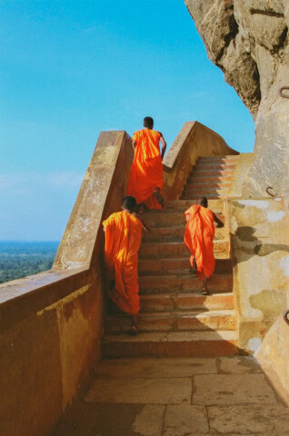 Three monks in orange robes ascend stone steps on a mountainside in Sri Lanka, against a backdrop of clear blue sky and distant landscape.
