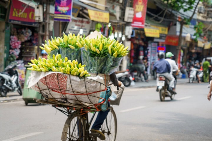 A vendor transports yellow flowers on a bicycle through a bustling street in Vietnam, filled with shops and motorbikes.