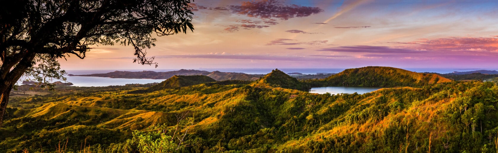 Panoramic view of rolling green hills, a large tree in the foreground, and a lake under a colorful sunset sky—scenery reminiscent of the best parks Madagascar has to offer.