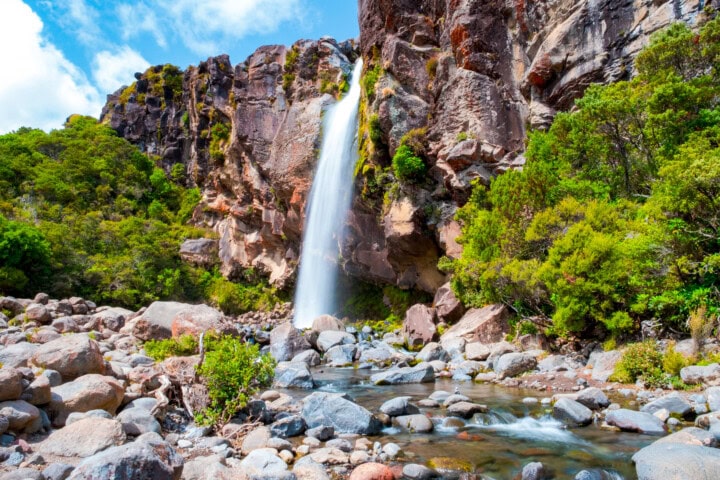 A waterfall cascades down a rocky cliff into a stream surrounded by lush green foliage and boulders under a partly cloudy New Zealand sky.