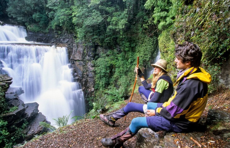 Two people in outdoor gear sit on a rocky ledge overlooking a multi-tiered waterfall surrounded by lush greenery, epitomizing Tasmania's stunning natural beauty. One holds a wooden walking stick while both look towards the falls, embodying the spirit of travel and tourism adventure.