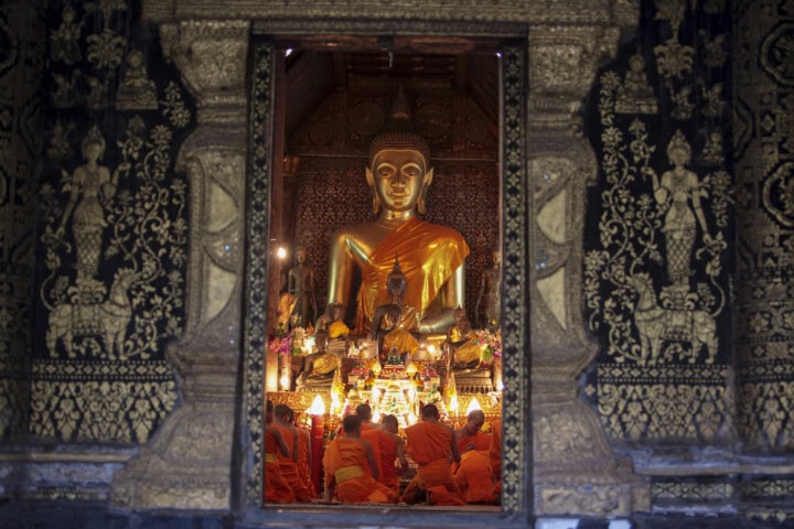 A group of monks in orange robes sit and face a large golden Buddha statue inside an ornately decorated temple in Laos.