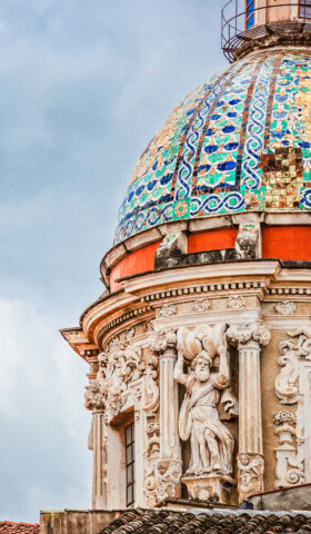 Close-up of an ornate dome featuring colorful mosaic patterns and intricate architectural details, including statues, against a partly cloudy sky—a stunning highlight of Italian tourism.