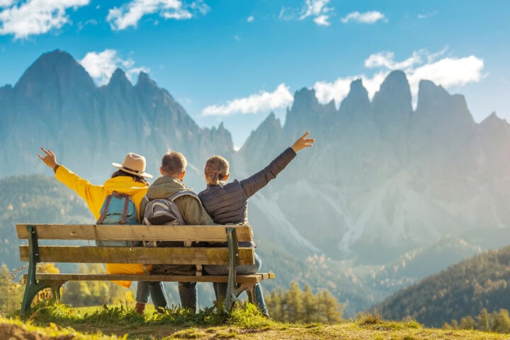 Three people sitting on a bench with their arms raised, facing a breathtaking mountainous landscape under Italy's clear sky, epitomizing the joy of travel and tourism.