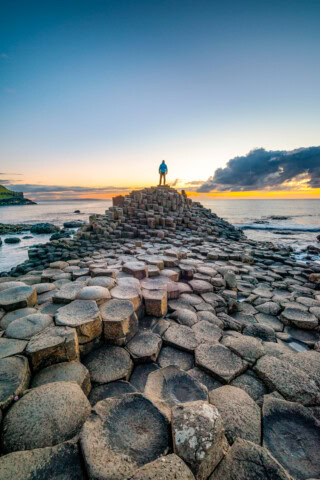 A person stands at the top of the Giant's Causeway in Ireland, a formation of hexagonal basalt columns, during sunset with the ocean and a cloudy sky in the background.
