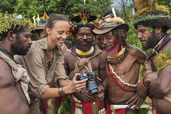 A woman shows a digital camera to five men dressed in traditional Papua New Guinea attire with ornamental headdresses and body paint, standing in an outdoor setting with trees in the background.