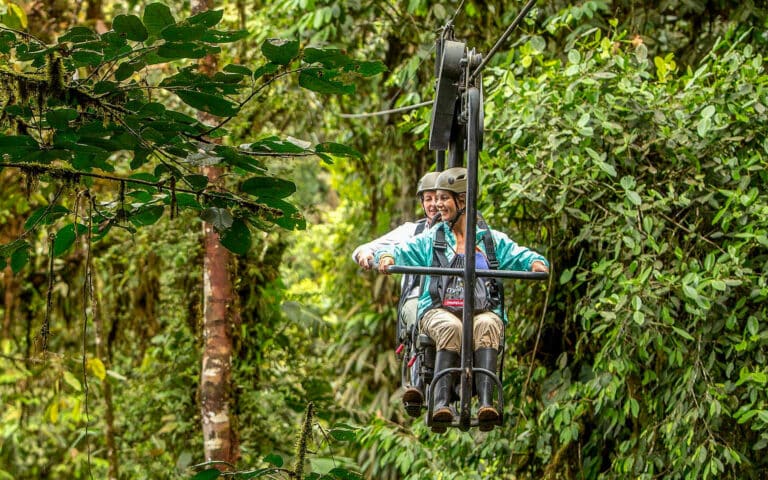 Two people riding a canopy cable car through a dense, green Ecuadorian forest, wearing helmets and outdoor gear.