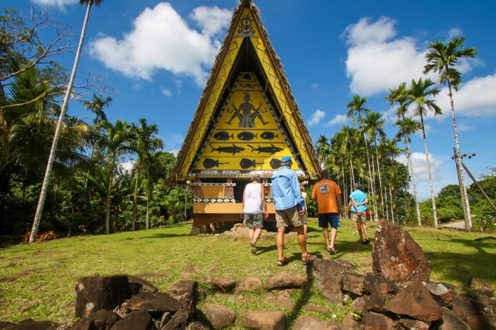 A group of people walk toward a traditionally decorated wooden structure with a triangular roof, surrounded by tropical trees and blue sky, in the enchanting travel destination of Palau.