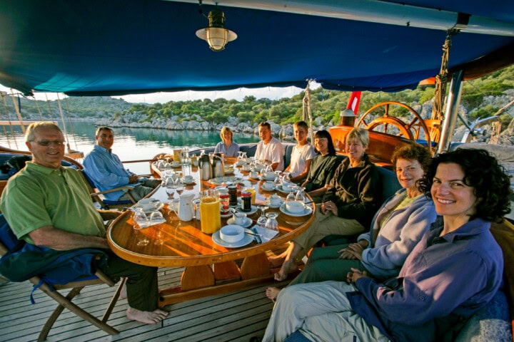 A group of eight people sit around a table on a boat, enjoying a meal with turkey at the center, all while taking in the serene water and greenery view in the background.