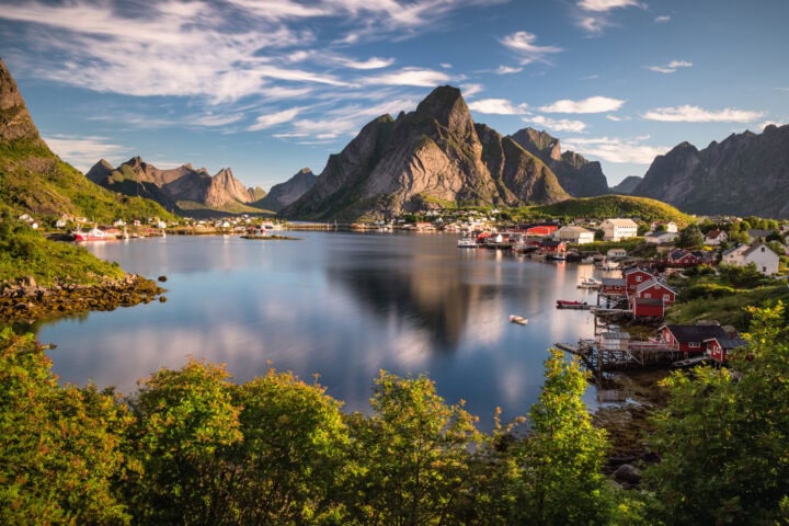 A calm fjord surrounded by steep mountains and a village with red and white houses, under a partly cloudy sky with greenery in the foreground.