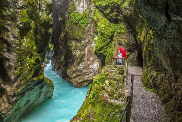 A person with a red umbrella stands on a path next to a turquoise river in a narrow, lush green gorge, capturing the essence of Slovenia's natural beauty, making it an ideal spot for travel and tourism.
