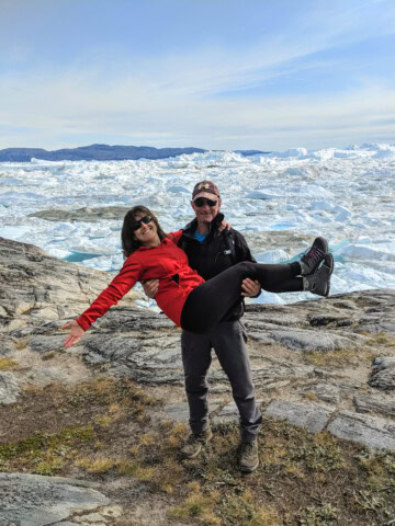 A man stands on Greenland's rocky terrain holding a woman in his arms, both smiling, with an icy landscape and mountains in the background on a clear day.