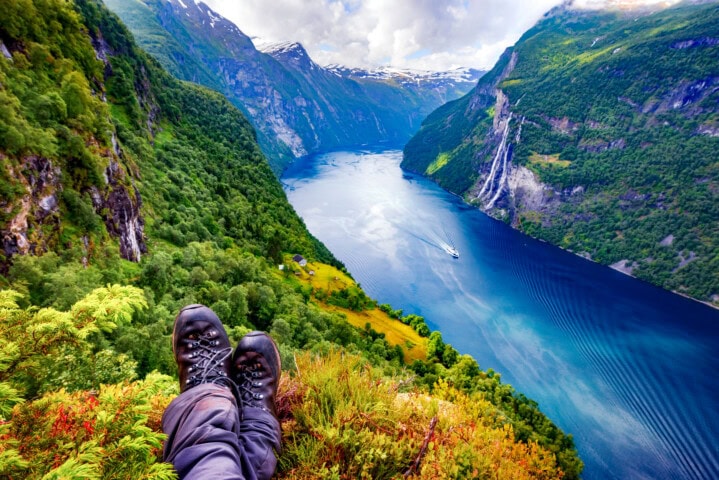 View from a cliff showing a pair of hiking boots overlooking Norway’s deep blue fjord, surrounded by green mountains, with a boat moving through the water and a waterfall visible on the right side.