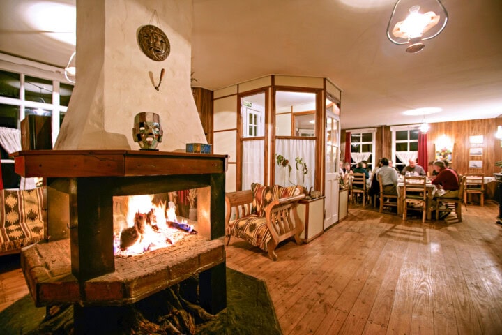 A warmly lit room with a cozy fireplace burning, wooden furniture, and people dining at tables in the background, evoking the charm of an Argentinean retreat.