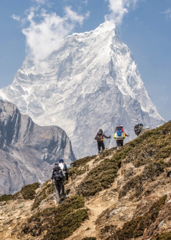 Hikers ascend a rugged trail with snow-capped mountains in the background under a clear sky, experiencing the breathtaking beauty that Nepal's tourism offers.