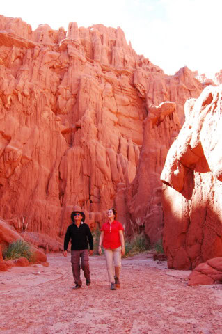 Two people walking through a canyon with red rock formations in Argentina, a clear sky overhead.