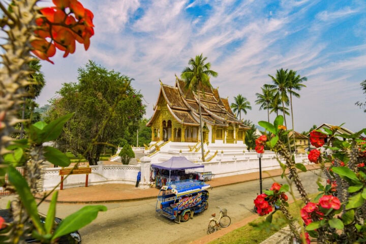 A vibrant tuk-tuk and bicycle pass by a traditional ornate temple surrounded by palm trees and flowers under a partly cloudy sky.