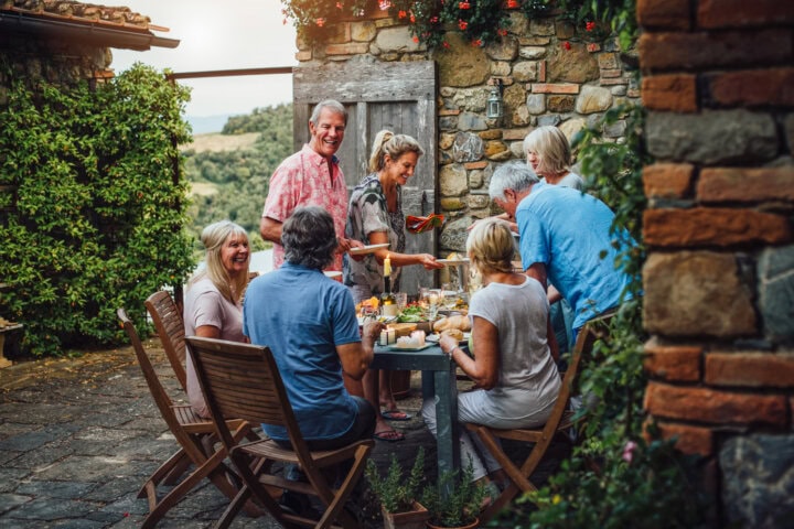 A group of mature friends are sitting around an outdoor dining table, eating and drinking. They are all talking happily and enjoying each others company in Tuscany, Italy.