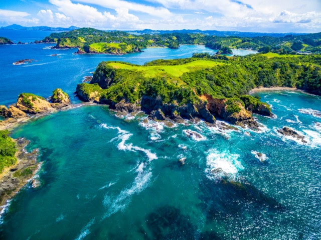 Aerial view of a coastal landscape in New Zealand featuring green cliffs, rocky shoreline, and turquoise waters with waves breaking. The horizon shows additional landmasses and a partly cloudy sky.