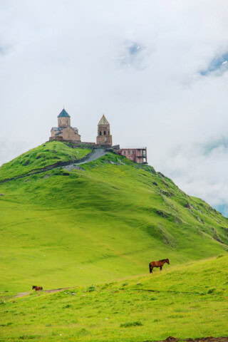 A historic stone church sits atop a green hill in Georgia, with two horses grazing in the foreground under a cloudy sky.