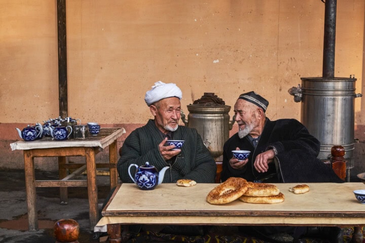 Two elderly men sit at an outdoor table with blue-patterned teaware and bread, engaging in conversation. Large metal urns are visible in the background, evoking images of traditional Uzbekistan gatherings.