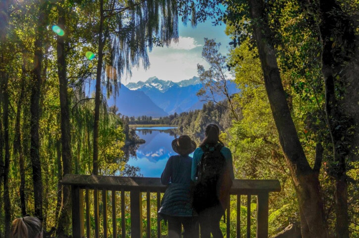 Two people stand on a wooden viewing platform overlooking a serene lake in New Zealand, surrounded by lush trees and mountains in the background under a clear sky.