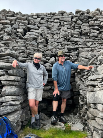 Two people stand in a rocky area, one leaning against a stone wall and the other placing a hand on a rock, capturing the rugged beauty of Ireland. Both are wearing casual outdoor attire. A backpack is visible on the left.