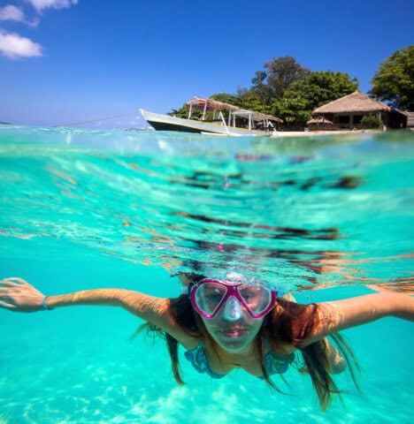 Woman snorkeling in the clear turquoise waters of Tonga, near boats and a thatched-roof hut on a sunny day in this stunning South Pacific island nation.