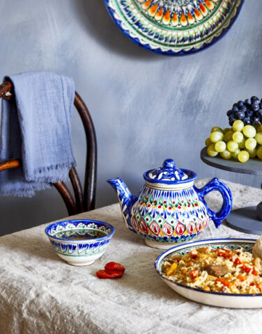 Table set with colorful ceramic teapot, bowl, and plate, featuring grapes on a tiered stand, a wooden chair with a folded blue cloth, and a wall decorated with a matching ceramic plate from Uzbekistan. This vibrant display captures the essence of Central Asia in every detail.