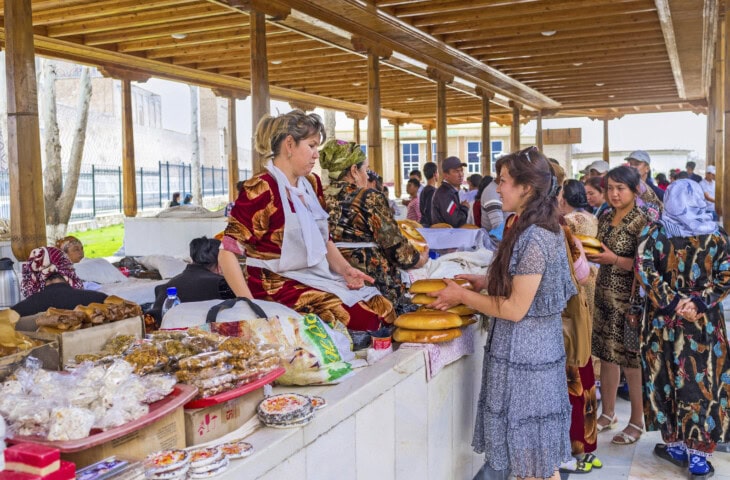 A woman buys bread from a vendor at a bustling market stall in Uzbekistan, filled with various food items. Other shoppers and vendors interact beneath the traditional wooden roof structure.