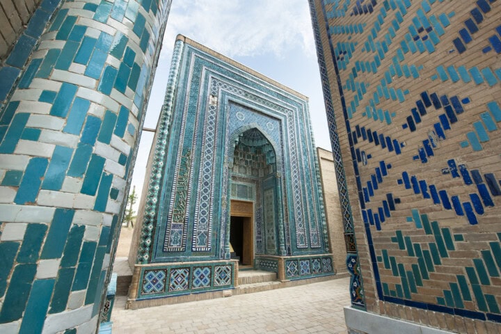 A narrow passageway between intricately decorated blue and beige tiled buildings, reminiscent of the Silk Road's historic routes through Uzbekistan, leading to a tall arched entrance.