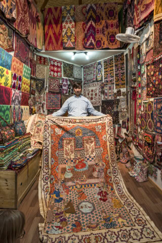 A person in a checked shirt displays a large, colorful, intricately patterned rug from Uzbekistan inside a shop filled with various fabrics and rugs hanging on the walls and displayed on shelves.