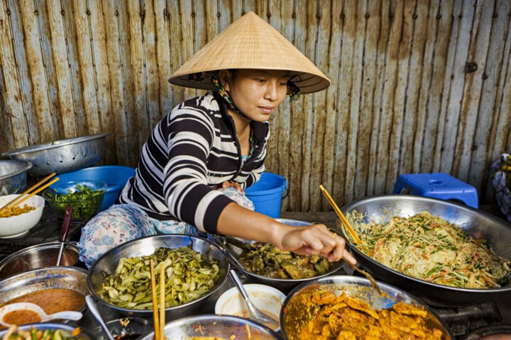 A person wearing a conical hat serves a variety of dishes from metal containers at a vibrant street food stall in Vietnam.