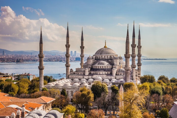 The image shows the Blue Mosque in Istanbul, Turkey, with its six minarets and cascading domes. It is surrounded by trees and urban landscape, with a body of water and distant cityscape in the background.