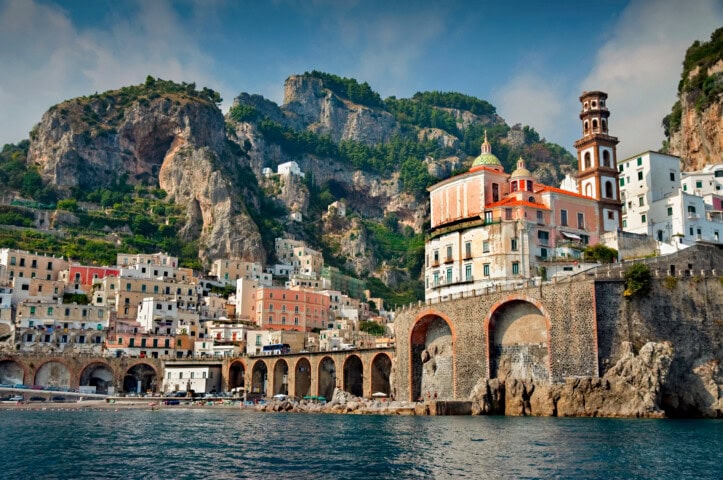 A coastal town in Italy with colorful buildings and a prominent tower positioned against a backdrop of rocky hills, adjacent to a calm waterfront.