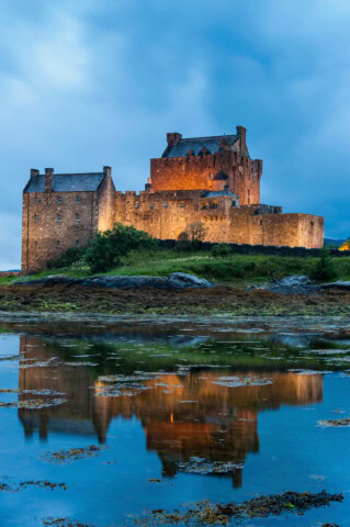 A large stone castle with turrets is reflected in a calm body of water. The sky is covered with dark clouds, and the surrounding green, rocky landscape evokes the essence of Scotland, making it an ideal travel destination for fans of historical tourism.