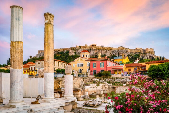 Ancient columns stand in the foreground, while colorful buildings and the Acropolis of Athens are visible in the background, under a vibrant Greece sky. Pink flowers are in the bottom right corner.