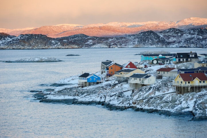 Coastal village in Greenland with colorful houses on a snow-covered rocky shoreline. Calm sea surrounds the area, with rugged hills in the background. Twilight casts a warm glow on the distant landscape.