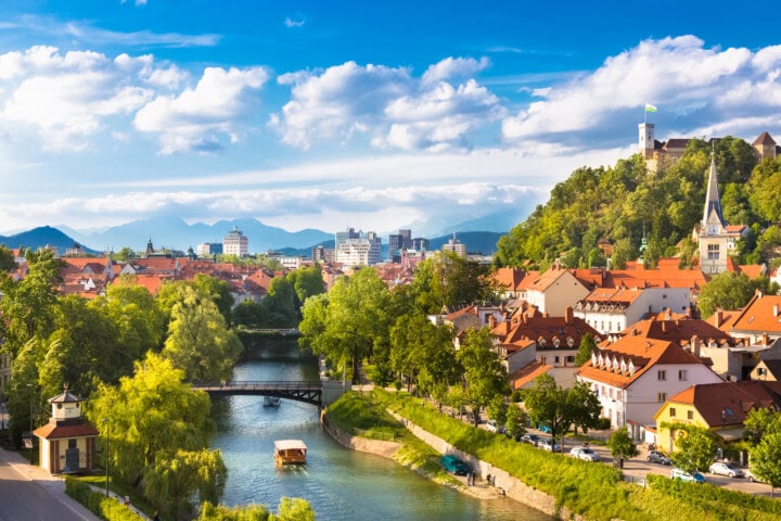 A riverside town in Slovenia with red-roofed buildings, a bridge over the river, lush greenery, a hilltop castle, and distant mountains under a partly cloudy sky.
