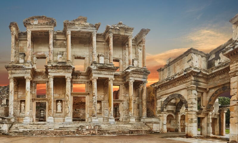 Ancient stone ruins of the Library of Celsus and Gate of Augustus in Ephesus, Turkey, are shown at sunset under a partly cloudy sky.