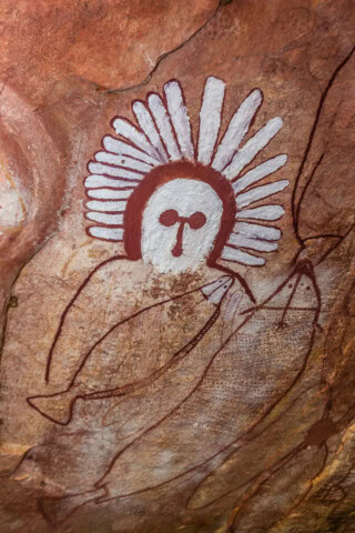 Ancient cave painting of a human-like figure with a white headdress and two fish, displayed on a red-brown rock surface in Australia.