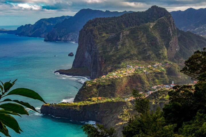 Coastal village on a steep, green cliff overlooking the ocean, with rugged mountains in the background under a cloudy sky.