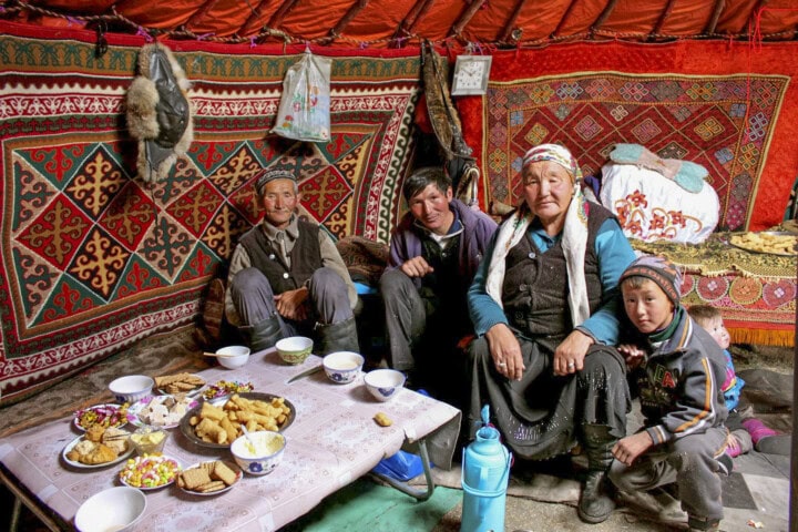A family sits inside a traditional Mongolian tent decorated with colorful textiles. A table in front of them is set with various dishes and snacks.