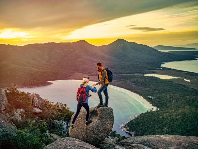 Two hikers with backpacks stand on a large rock overlooking a coastal bay and mountain landscape at sunset, with one helping the other balance.