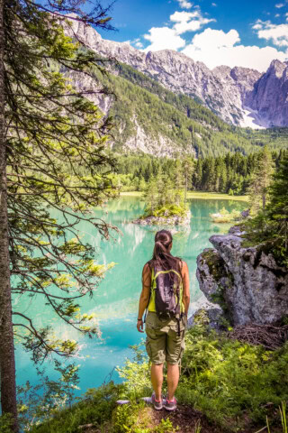 A person with a backpack stands on a grassy ledge, overlooking a turquoise lake with surrounding trees and mountains under a partly cloudy sky, reminiscent of the picturesque landscapes found in Italy.