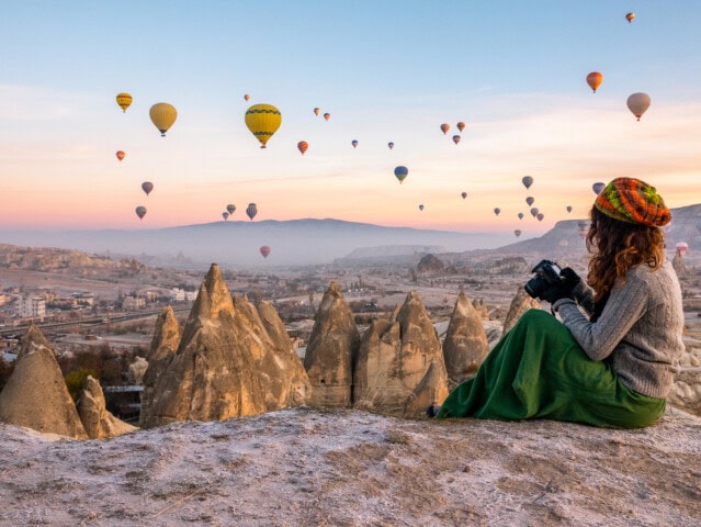 A person with a camera sits on a rocky ledge in Turkey, overlooking a landscape with numerous hot air balloons in the sky during sunrise or sunset.