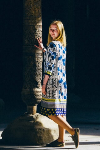 A person stands beside an intricately carved pillar in Uzbekistan, wearing a blue and white patterned dress and brown shoes, smiling at the camera.