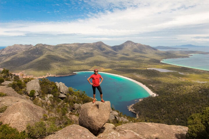 Person stands on a rock overlooking a green hilly landscape and a clear, crescent-shaped bay under a partly cloudy sky, embracing nature's beauty while considering their next travel adventure to Tasmania.